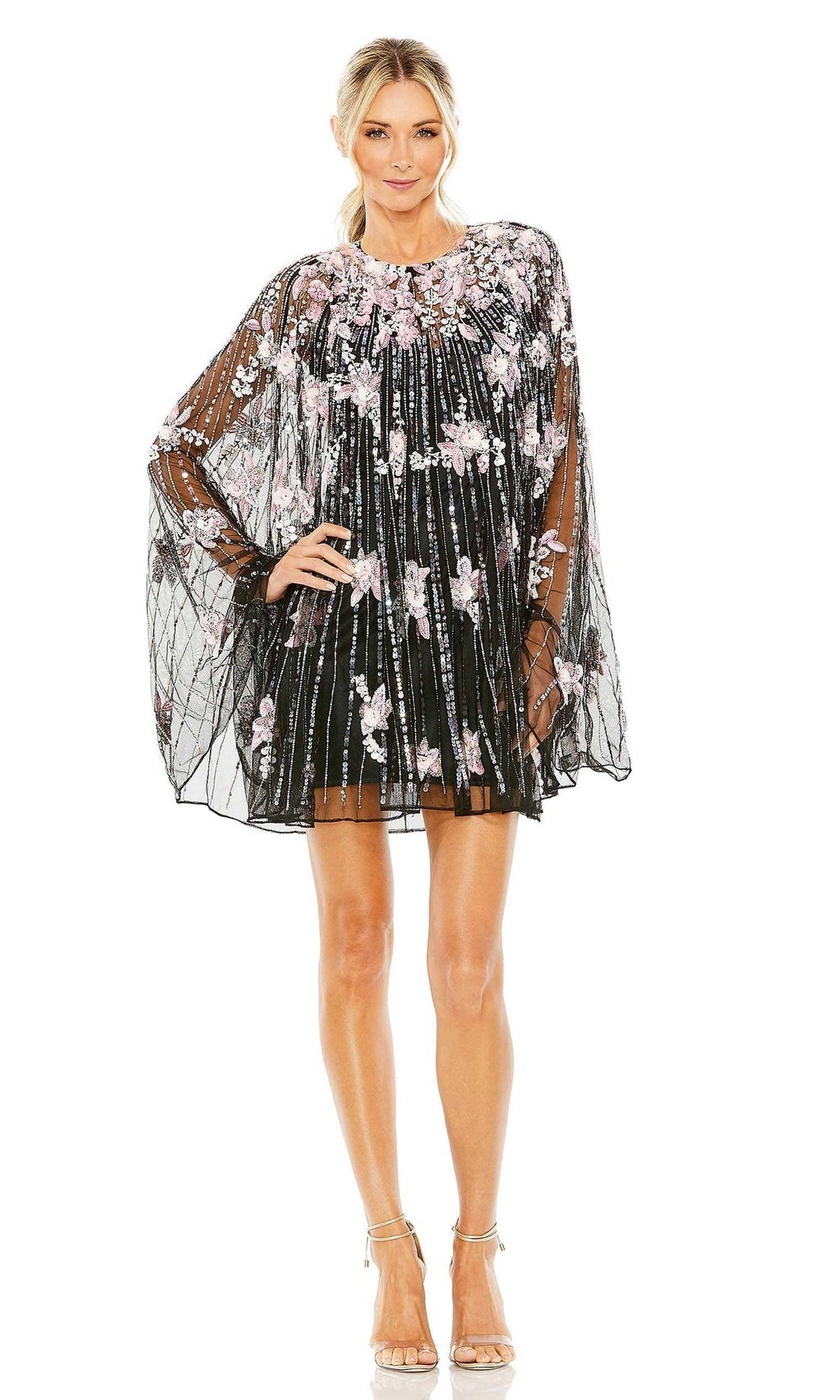Model wears the Beaded-Cape Short Hoco Dress (Mac Duggal 5917)—a sheer, black, floral-embroidered, loose-fit dress with a beaded cape—paired with strappy heels against a white background.